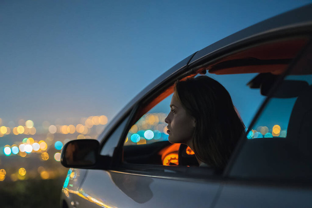 Ta žena sedí v autě na pozadí osvětlujícím město. večerní čas - Fotografie, Obrázek