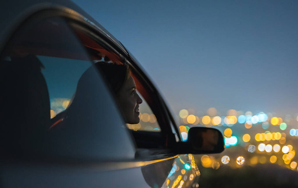 La femme heureuse s'assied dans une voiture sur le fond des lumières de la ville. soir heure de nuit - Photo, image