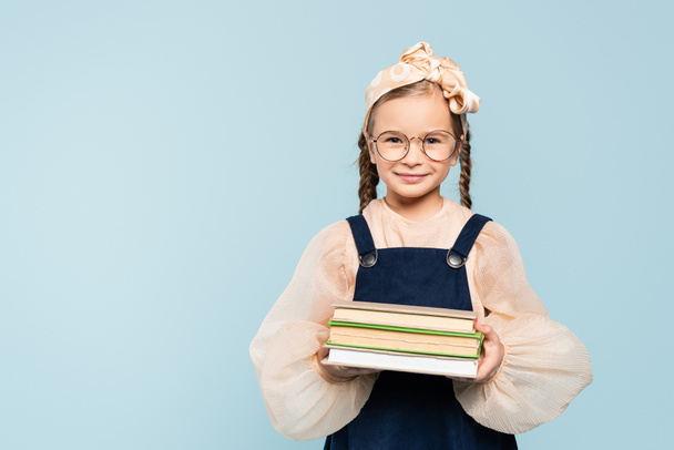smart kid in glasses smiling while holding books isolated on blue - Foto, Imagem