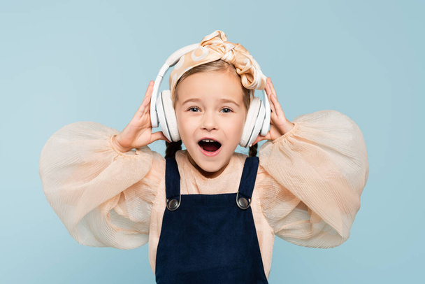 amazed kid in headband with bow and wireless headphones listening music isolated on blue  - Photo, Image