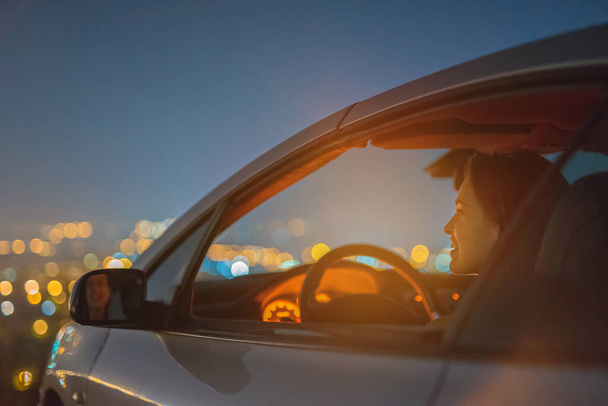 Mutlu kadın şehrin aydınlık arka planında bir arabada oturuyor. Akşam vakti. - Fotoğraf, Görsel