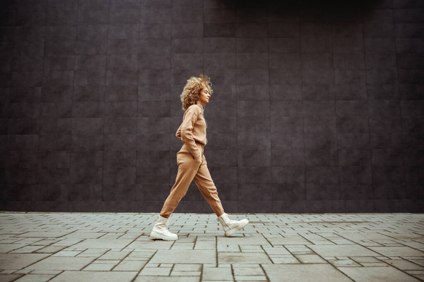 Πλευρική άποψη του κοριτσιού παγίδα σε tracksuit περπάτημα με τα χέρια στις τσέπες της και ακούγοντας μουσική. - Φωτογραφία, εικόνα