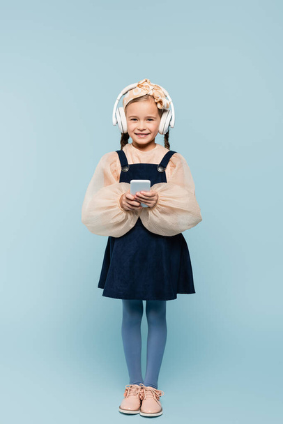 full length of cheerful kid in headband with bow and wireless headphones using smartphone isolated on blue  - Foto, Imagem