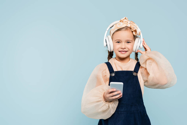 cheerful kid in headband with bow adjusting wireless headphones while holding smartphone isolated on blue  - Foto, immagini