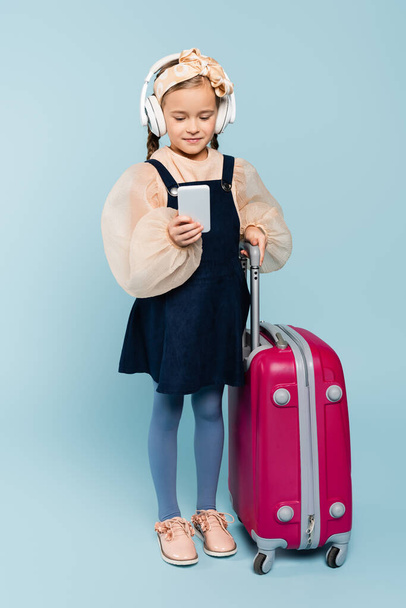 full length of little girl in wireless headphones using smartphone while standing with baggage on blue  - Foto, immagini
