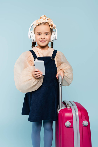 niña feliz en auriculares inalámbricos utilizando el teléfono inteligente mientras está de pie con el equipaje en azul  - Foto, Imagen