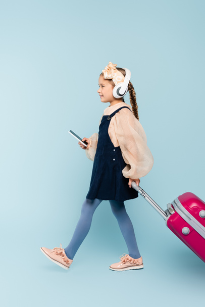toute la longueur de la petite fille dans les écouteurs sans fil tenant smartphone tout en marchant avec des bagages sur bleu  - Photo, image