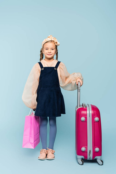 full length of happy little girl holding shopping bag and standing with baggage on blue  - Foto, Imagem