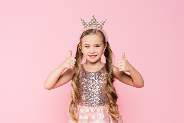 χαρούμενο κοριτσάκι με φόρεμα και στέμμα που δείχνει τους αντίχειρες επάνω απομονωμένο σε ροζ  - Φωτογραφία, εικόνα