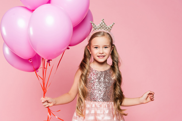 joyeuse petite fille en robe et couronne tenant des ballons isolés sur rose - Photo, image