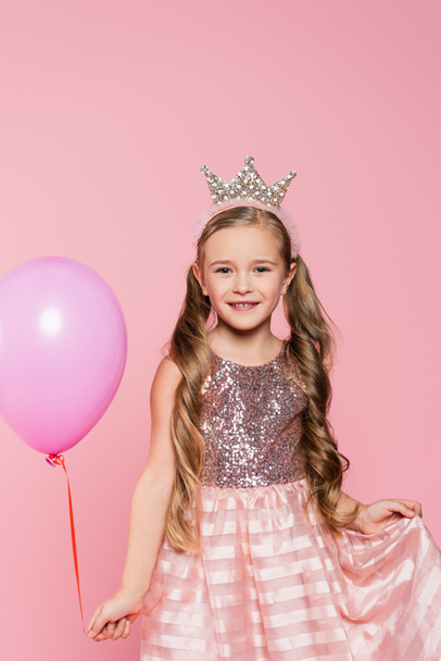 joyful little girl in dress and crown holding balloon isolated on pink - 写真・画像