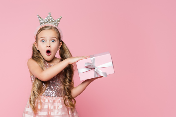 amazed little girl in crown holding wrapped present isolated on pink  - 写真・画像