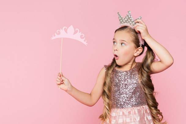 shocked little girl in dress holding paper crown on stick isolated on pink  - Photo, Image