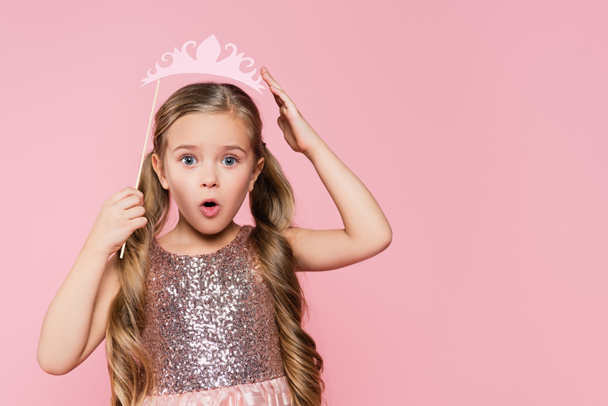 verrast klein meisje in jurk houden papier kroon op stok boven hoofd geïsoleerd op roze  - Foto, afbeelding