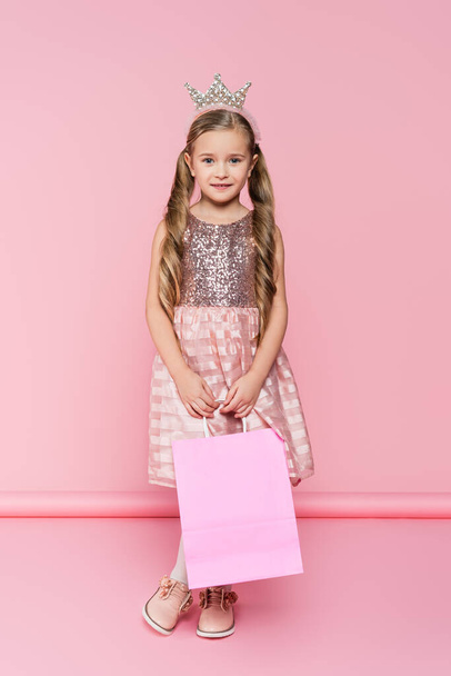 full length of cheerful little girl in dress and crown holding shopping bag on pink  - Fotoğraf, Görsel