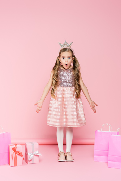 full length of shocked little girl in crown standing near presents and shopping bags on pink  - Фото, изображение