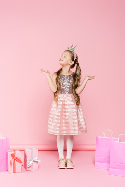 full length of pleased little girl in crown with closed eyes standing near presents and shopping bags on pink  - Fotografie, Obrázek