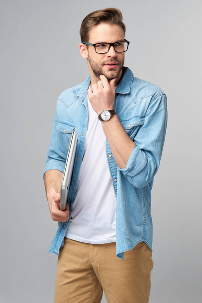 Concentrated young bearded man wearing glasses dressed in jeans shirt holding laptop isolated over grey studio background - Zdjęcie, obraz