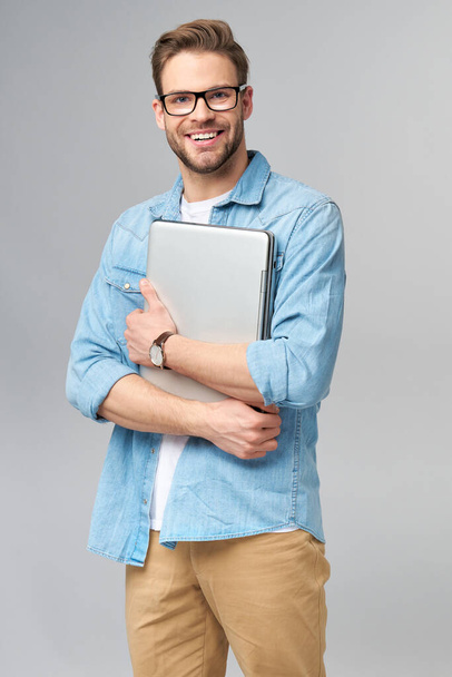 Concentrated young bearded man wearing glasses dressed in jeans shirt holding laptop isolated over grey studio background - Foto, imagen