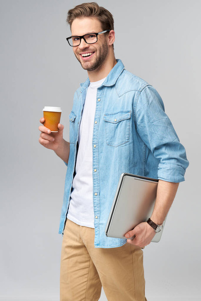 Concentrated young bearded man wearing glasses dressed in jeans shirt holding laptop and cup of coffee to go isolated over grey studio background - Фото, изображение
