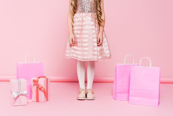 bijgesneden weergave van klein meisje in jurk staan in de buurt van cadeautjes en boodschappentassen op roze  - Foto, afbeelding