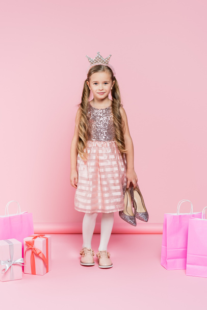повна довжина щасливої маленької дівчинки в короні тримає підбори біля подарунків і сумки для покупок на рожевому
  - Фото, зображення