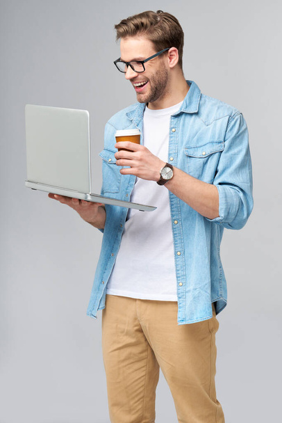 Concentrated young bearded man wearing glasses dressed in jeans shirt holding laptop and cup of coffee to go isolated over grey studio background - Foto, Imagem