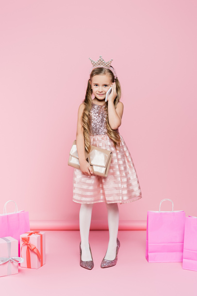 full length of happy little girl in crown standing on heels and talking on smartphone near presents and shopping bags on pink  - Foto, afbeelding
