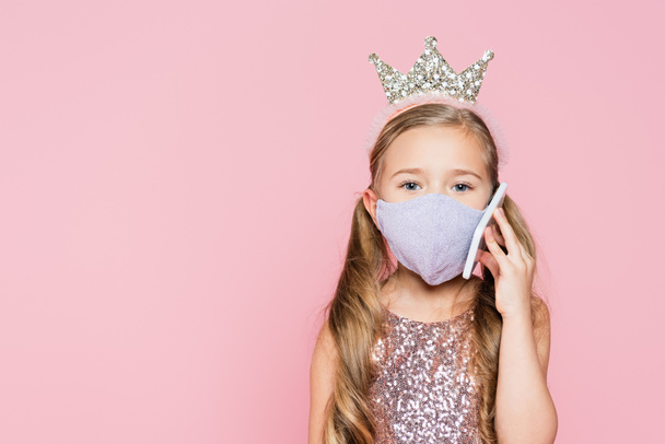 Mädchen in Krone und medizinischer Maske spricht auf Smartphone isoliert auf rosa  - Foto, Bild