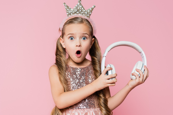 wstrząśnięta dziewczynka w koronie trzyma słuchawki bezprzewodowe izolowane na różowy - Zdjęcie, obraz