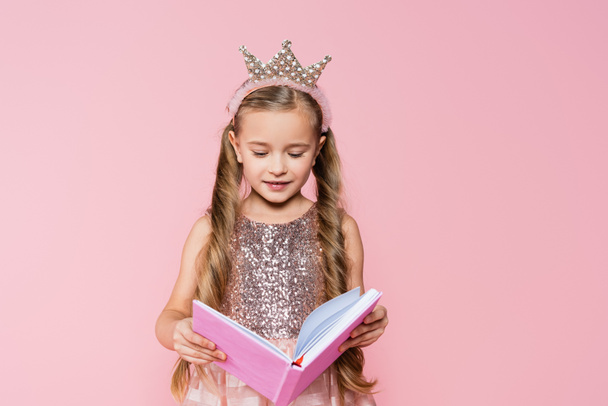 happy little girl in crown reading book isolated on pink - Foto, imagen