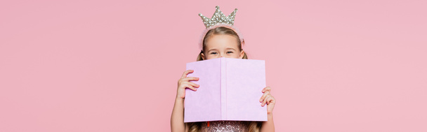 κοριτσάκι με κορώνα που καλύπτει το πρόσωπο με το βιβλίο απομονωμένο σε ροζ, banner - Φωτογραφία, εικόνα