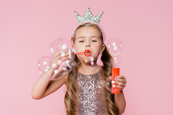niña en corona soplando burbujas de jabón en primer plano borroso aislado en rosa  - Foto, Imagen