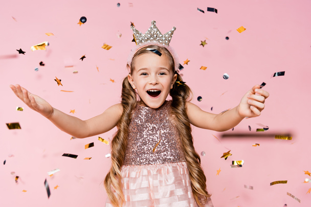 asombrada niña en corona con las manos extendidas cerca de caer confeti en rosa  - Foto, Imagen