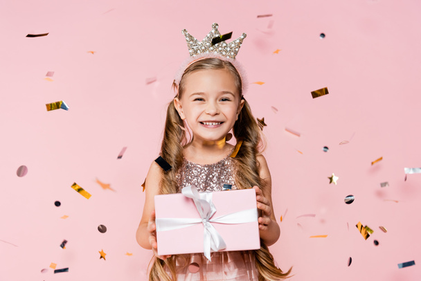 heureuse petite fille en couronne tenant présent près de tomber confettis sur rose  - Photo, image