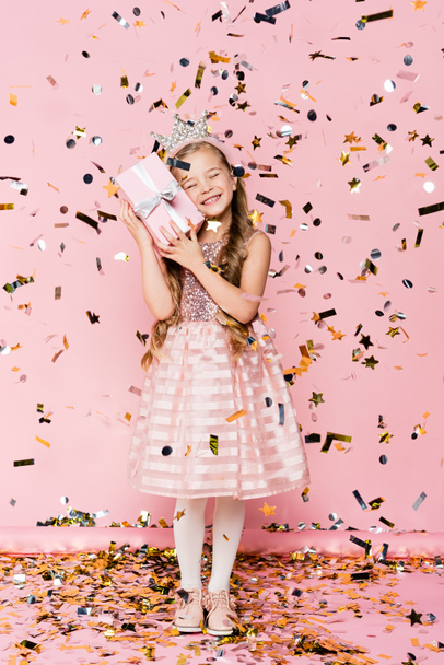full length of happy little girl in crown holding present near falling confetti on pink  - Photo, Image