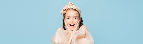 amazed kid in headband with bow touching cheeks isolated on blue, banner - Foto, afbeelding
