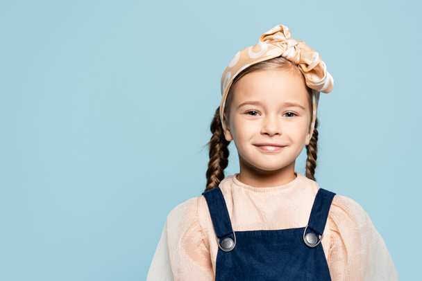 fröhliches Kind mit Stirnband und Schleife, das isoliert auf blau in die Kamera blickt - Foto, Bild