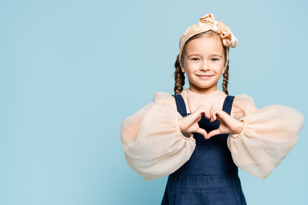 cheerful kid in headband with bow looking at camera and showing heart with hands isolated on blue - Foto, Imagen