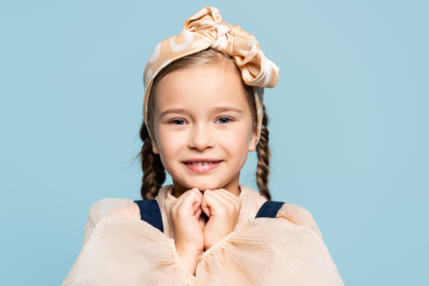 positive kid in headband with bow looking at camera isolated on blue - Foto, afbeelding