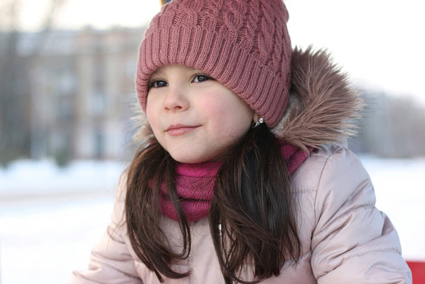 Kis mosolygós, fehér lány hosszú, sötét hajjal, kötött kalapban, hóban és homályos épületekben. - Fotó, kép