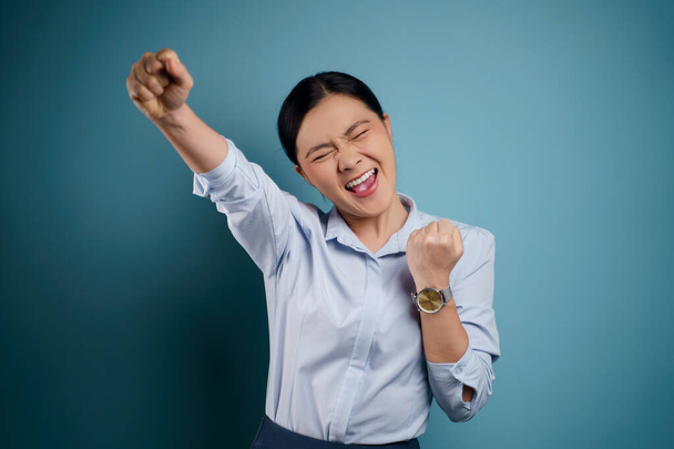 Азиатка счастлива стоя показывая свой кулак сделать победный жест изолирован на синем фоне. - Фото, изображение
