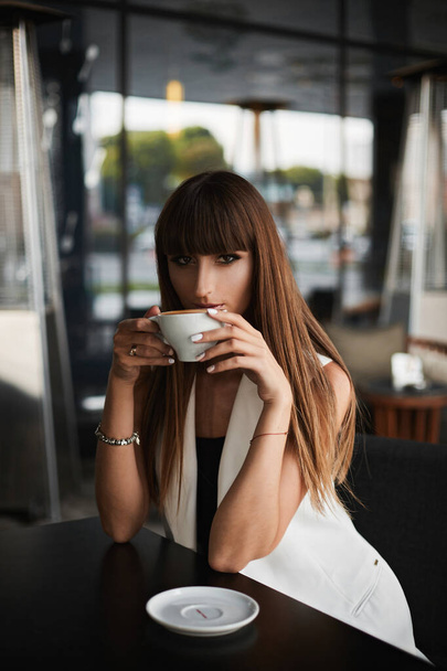 Young mixed race model woman with a seductive look and bright makeup drinks tasty cappuccino in an empty cafe - Фото, зображення