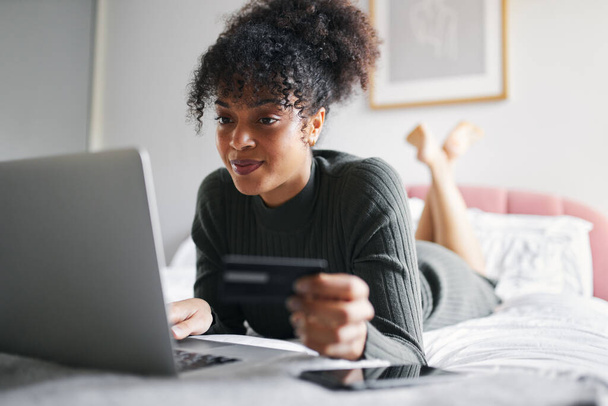 Молодая женщина дома лежит на кровати с ноутбуком с помощью кредитной карты, чтобы совершать покупки в Интернете - Фото, изображение