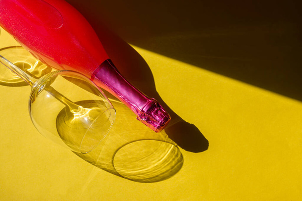Feestelijk partijconcept. Glas en fles roze champagne op een gele achtergrond. Zonlicht onthult de vaste vormen van glas en flesschaduwen. Hard licht, harde schaduwen. Vlakke plaat. - Foto, afbeelding