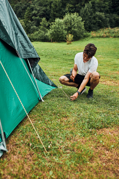 Young man putting up a tent on camping during summer vacation trip. Teenager putting the stakes into grassy ground using hammer - Foto, afbeelding