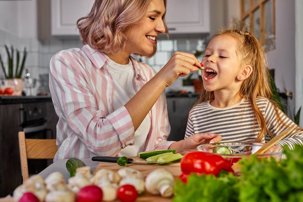 cute mom and child girl tasting fresh fruits while preparing salad for dinner - Photo, Image