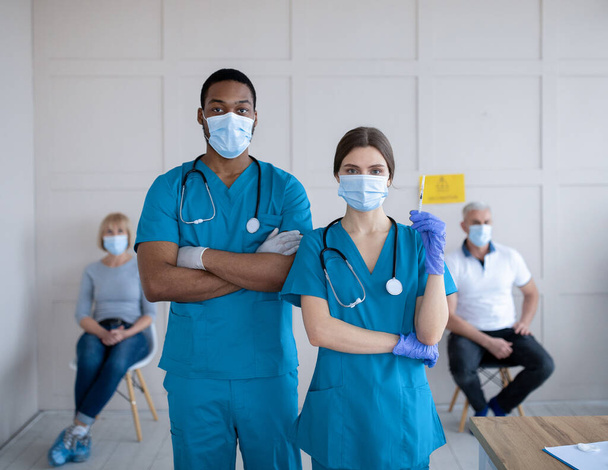 Male and female doctors wearing uniforms and face masks, holding syringe with coronavirus vaccine at clinic - Φωτογραφία, εικόνα