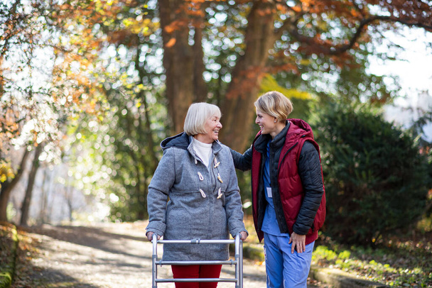 Senior woman with walking frame and caregiver outdoors on a walk in park, coronavirus concept. - Photo, Image