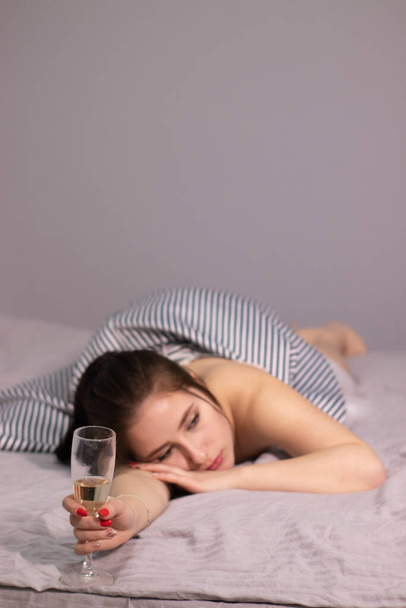 smutna atrakcyjna brunetka w wieczorowej sukni koktajlowej ze szklanką szampana na łóżku w szarej sypialni. Samotna impreza, samotność. przestrzeń kopiowania  - Zdjęcie, obraz
